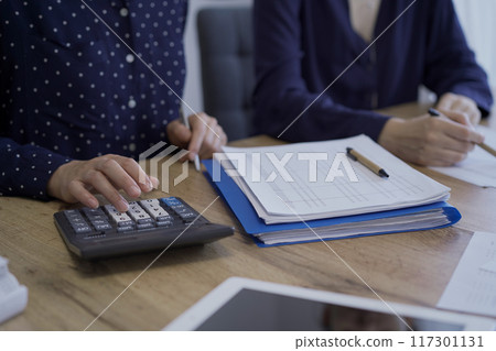 Accountants or auditors reviewing financial documents while sitting at the wooden desk in office. Professionals analyzing paper reports and using calculators, side view. Audit and taxes 117301131