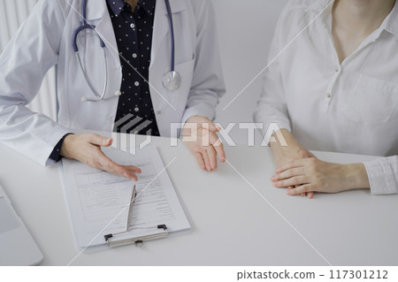 Doctor and a patient. The female physician, wearing a white medical coat over a dark blue dotted blouse, using clipboard and making notes during a consultation in the clinic, close up. Medicine Doctor and a patient. The female physician, wearing a white medical coat over a dark blue dotted blouse, using clipboard and making notes during a consultation in the clinic, close up. Medicine 117301212