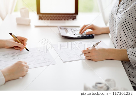 Two accountants using a calculator and laptop computer for counting taxes or revenue balance while rolls of receipts are waiting to be calculating. Business audit and taxes concepts 117301222