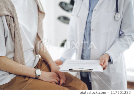 Doctor and patient in clinic. Friendly physician explaining something to young woman while keeping a clipboard with medical records. Medicine concept Doctor and patient in clinic. Friendly physician explaining something to young woman while keeping a clipboard with medical records. Medicine concept 117301223
