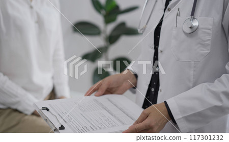 Doctor and a patient discussing something. The female physician, wearing a white medical coat is keeping clipboard with a medical notes besides a young woman during a consultation in the clinic Doctor and a patient discussing something. The female physician, wearing a white medical coat is keeping clipboard with a medical notes besides a young woman during a consultation in the clinic 117301232