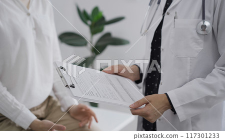 Doctor and a patient discussing something. The female physician, wearing a white medical coat is keeping clipboard with a medical notes besides a young woman during a consultation in the clinic Doctor and a patient discussing something. The female physician, wearing a white medical coat is keeping clipboard with a medical notes besides a young woman during a consultation in the clinic 117301233