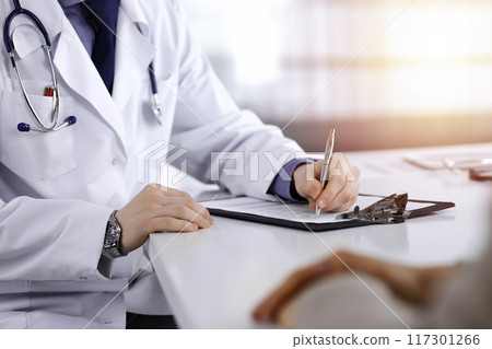 Unknown male doctor and patient woman discussing something while sittingin a darkened clinic and using clipboard, glare of light on the background Unknown male doctor and patient woman discussing something while sittingin a darkened clinic and using clipboard, glare of light on the background 117301266