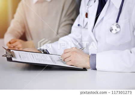 Unknown male doctor and patient woman discussing something while sittingin a darkened clinic and using clipboard, glare of light on the background Unknown male doctor and patient woman discussing something while sittingin a darkened clinic and using clipboard, glare of light on the background 117301273