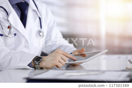 Unknown male doctor sitting and working with tablet computer iin a darkened clinic, glare of light on the background, close-up of hands Unknown male doctor sitting and working with tablet computer iin a darkened clinic, glare of light on the background, close-up of hands 117301389
