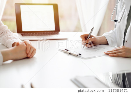 Doctor and patient sitting near each other at the white desk in clinic. Female physician is listening filling up a records form. Medicine concept 117301471
