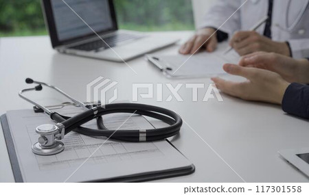 Stethoscope over a clipboard with medical papers is lying on the table near doctor and patient discussing health while sitting in clinic. Medicine and science Stethoscope over a clipboard with medical papers is lying on the table near doctor and patient discussing health while sitting in clinic. Medicine and science 117301558