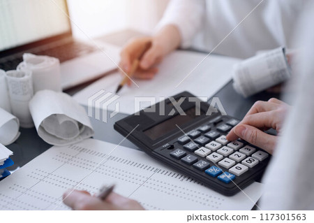 Two accountants use a calculator and laptop computer for counting taxes or revenue balance. Business, audit, and taxes concepts Two accountants use a calculator and laptop computer for counting taxes or revenue balance. Business, audit, and taxes concepts 117301563