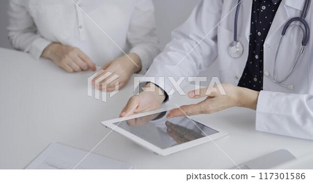 Doctor and a patient. The female physician, wearing a white medical coat over a dark blue dotted blouse, using tablet computer during a consultation in the clinic. Medicine concept Doctor and a patient. The female physician, wearing a white medical coat over a dark blue dotted blouse, using tablet computer during a consultation in the clinic. Medicine concept 117301586