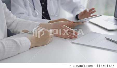 Doctor and a patient. The female physician, wearing a white medical coat over a dark blue dotted blouse, using tablet computer during a consultation in the clinic. Medicine concept Doctor and a patient. The female physician, wearing a white medical coat over a dark blue dotted blouse, using tablet computer during a consultation in the clinic. Medicine concept 117301587