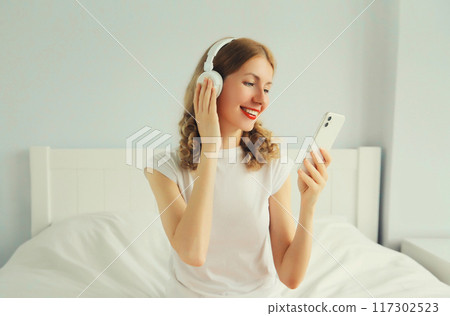 Happy smiling young woman listening to music with headphones looking at phone on bed at home 117302523