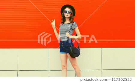 Beautiful woman model posing wearing black hat, shorts and handbag in the city on orange background 117302533