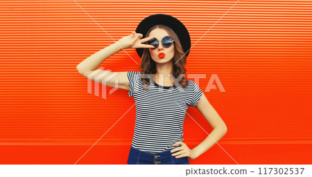Portrait of beautiful young woman blowing kiss wearing black round hat on orange background 117302537