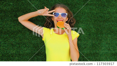 Portrait of happy smiling young woman with big burger fast food lying on the grass in summer park Portrait of happy smiling young woman with big burger fast food lying on the grass in summer park 117303917
