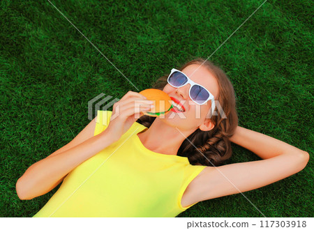 Portrait of happy smiling young woman with big burger fast food lying on the grass in summer park Portrait of happy smiling young woman with big burger fast food lying on the grass in summer park 117303918