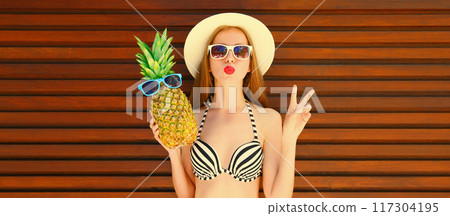 Summer portrait of happy young woman with pineapple blowing kiss in summer straw hat Summer portrait of happy young woman with pineapple blowing kiss in summer straw hat 117304195