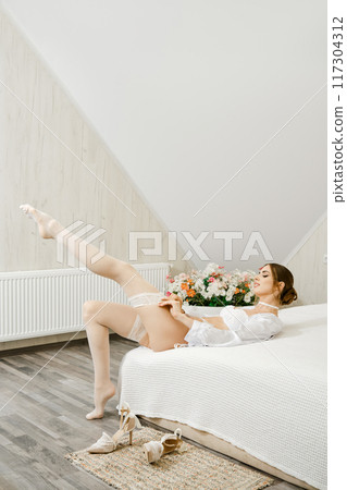 Playful woman laying down on a white bed and putting on white stockings 117304312