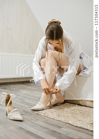 Young woman fastens the buckle on her shoes while sitting on the bed Young woman fastens the buckle on her shoes while sitting on the bed 117304315