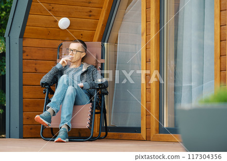 Middle-aged man smoking a cigar while sitting on the terrace of a country house 117304356