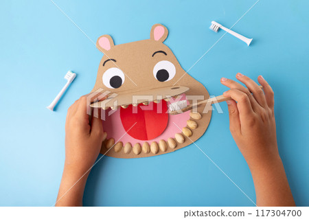 Paper craft portraying a hippo puppet, crafted for kids using cardboard. wide open mouth displays pistachio teeth, with a hand holding a toothbrush, aimed at educating children about dental hygiene 117304700