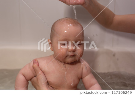 A calm baby enjoying a gentle bath, water streaming softly over their peaceful face. 117306249
