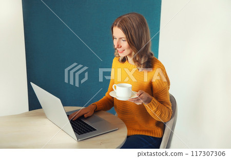 Modern young woman working with laptop drinking coffee sitting at the desk in the room at home Modern young woman working with laptop drinking coffee sitting at the desk in the room at home 117307306