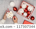 Red and white Christmas baubles in a box flat lay. Merry Christmas and Happy New Year seasons greetings card. Copy space. Festive full frame. Hands holding hot dink cup 117307344