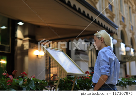 A woman is thoroughly reviewing the menu while seated at an elegant outdoor cafe venue 117307975