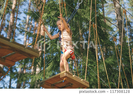 A brave strong Child walks along the suspended slats of a rope track at a height among pine trees 117308128