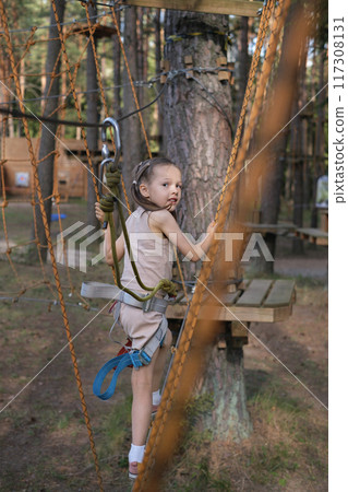 A preschool-age girl climbs a rope suspension bridge in a wooded area 117308131