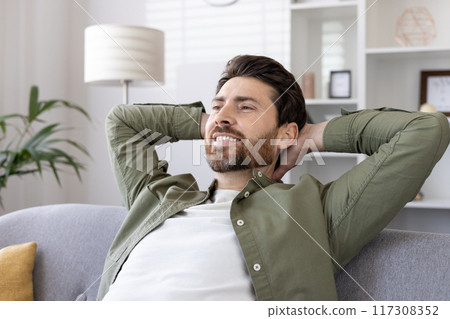 Relaxed man sitting on sofa with hands behind head, enjoying leisure time at home. Man smiling while resting and feeling stress-free indoors. 117308352