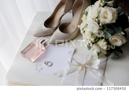 Bride's wedding essentials: heels, bouquet, rings, perfume, and invitation on a white table Bride's wedding essentials: heels, bouquet, rings, perfume, and invitation on a white table 117308450
