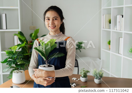 Portrait of a smiling young woman holding potted plant in the house and taking care of the greenery of the house 117311042