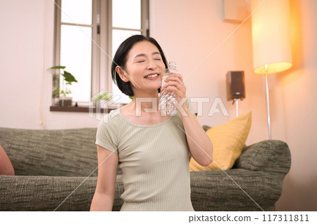 Middle-aged woman in sportswear drinking bottled water in the living room Middle-aged woman in sportswear drinking bottled water in the living room 117311811