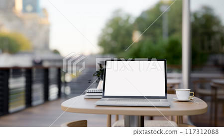 A laptop computer with a white-screen mockup on a table on a coffee shop outdoor terrace or balcony. 117312088