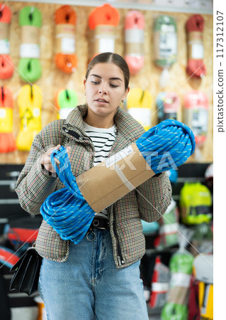 Young girl in winter clothes carefully selects climbing rope for winter hike in sports equipment store 117312107