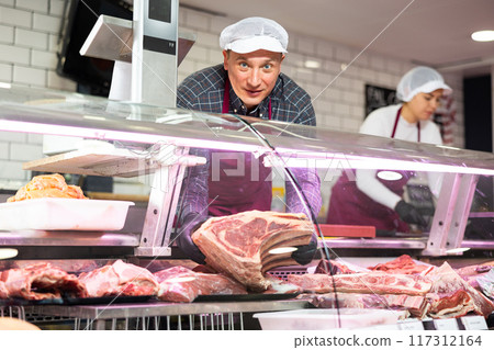 Diligent male butcher offering to sell big piece of beef beef in butcher shop 117312164
