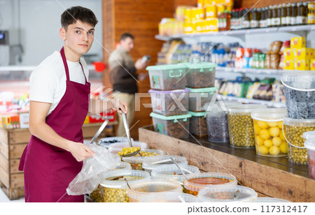 Friendly salesman in an apron offers to buy pickled green olives in grocery supermarket 117312417
