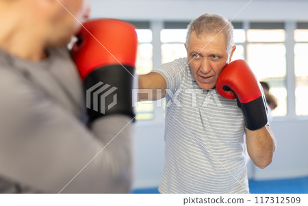 Boxing fight - elderly man learning to box in sparring with trainer in gym Boxing fight - elderly man learning to box in sparring with trainer in gym 117312509
