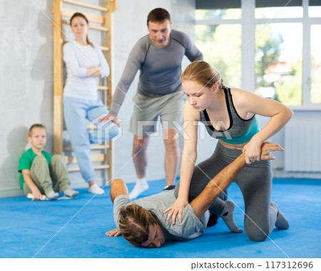 Focused girl in sport club during training, sparring and practicing technique of grabbing arm and lowering partner to mat with help of trainer Focused girl in sport club during training, sparring and practicing technique of grabbing arm and lowering partner to mat with help of trainer 117312696