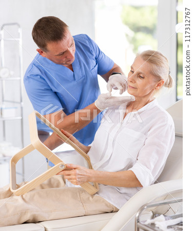 Mature woman sitting with mirror and discussing with male doctor for rejuvenation procedure Mature woman sitting with mirror and discussing with male doctor for rejuvenation procedure 117312767