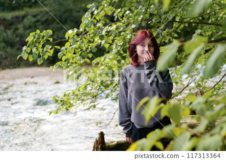 Rebellious portrait of a modern teen with dyed hair and facial piercings, set against the lush, natural backdrop of a forested river landscape Rebellious portrait of a modern teen with dyed hair and facial piercings, set against the lush, natural backdrop of a forested river landscape 117313364