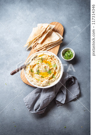 Bowl of creamy hummus garnished with olive oil and herbs is served with pita bread on a wooden board. A small bowl of garnish is placed beside the bread on a slate surface Bowl of creamy hummus garnished with olive oil and herbs is served with pita bread on a wooden board. A small bowl of garnish is placed beside the bread on a slate surface 117314346