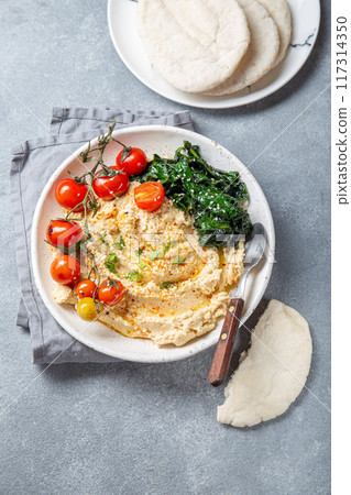 A delicious plate of creamy hummus garnished with fresh tomatoes, leafy greens, and served with pita bread 117314350