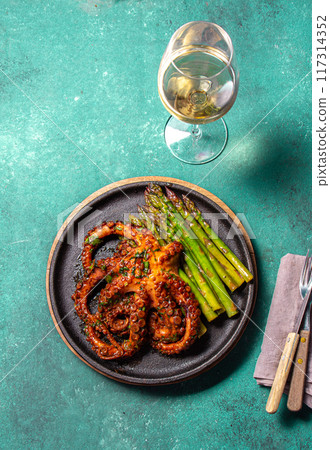 Grilled octopus with asparagus served on a black plate, highlighting vibrant colors and textures against a turquoise background. Grilled octopus with asparagus served on a black plate, highlighting vibrant colors and textures against a turquoise background. 117314352