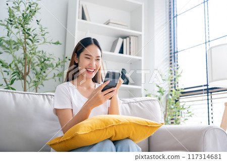 Happy young asian woman relax on comfortable couch at home texting messaging on smartphone 117314681