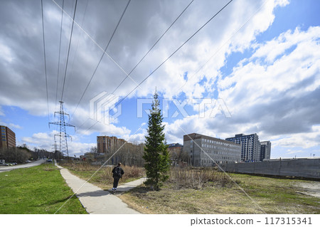 Cloudy sky over a high-voltage power line, buildings and a boy on a scooter Cloudy sky over a high-voltage power line, buildings and a boy on a scooter 117315341