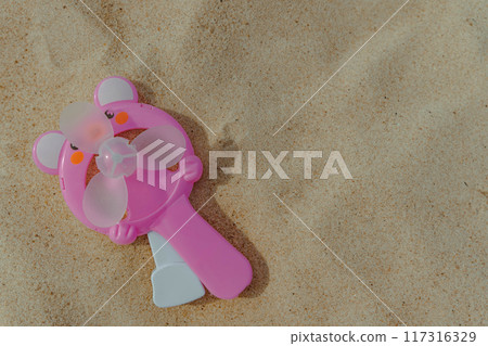 A pink handheld fan lies on soft sand at the beach 117316329