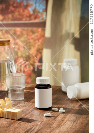 Creative scene for advertising with pharmacy theme. A medicine bottle with white label displayed on wooden table with glass cup, bottles and daily medicine box. Behind is with with autumn landscape 117316770