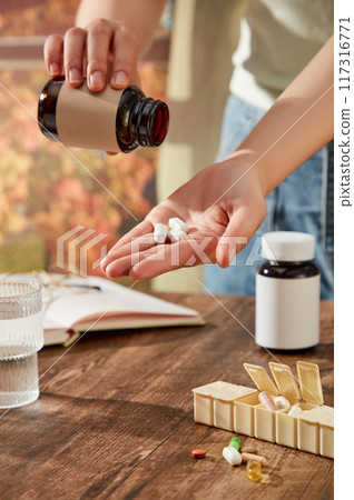 Scene with lifestyle drug advertisement. A woman's hand is pouring medicine in preparation for drinking. Medicine bottles without label on wooden table with glass water cup 117316771